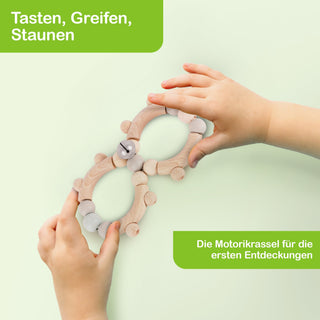 Zwei Kinderhände halten ein hölzernes Motorikspielzeug, das aus zwei ovalen Ringen besteht, die durch bewegliche Verbindungen und kleine Kugeln miteinander verbunden sind. Oben links im Bild steht in einem grünen Feld: ‚Tasten, Greifen, Staunen‘. Unten rechts befindet sich ein weiteres grünes Feld mit der Aufschrift: ‚Die Motorikrassel für die ersten Entdeckungen‘.