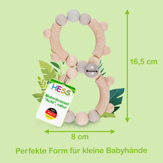 Motorikrassel in Form einer Acht aus naturfarbenem Holz, 16,5 cm hoch und 8 cm breit. Ein Etikett an der Rassel trägt die Aufschrift ‚HESS Motorikrassel ‚Acht‘, natur‘ und ‚100% Made in Germany‘. Im Hintergrund sind grüne Blätter zu sehen. Am unteren Bildrand steht der Text: ‚Perfekte Form für kleine Babyhände‘.