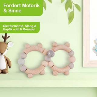 Auf einem hellgrünen Hintergrund steht ein Regal mit einem kleinen Spielzeug-Reh auf der linken Seite und zwei hölzernen Babyspielzeugen in Ringform mit Perlen und einer Glocke in der Mitte. Darüber befinden sich zwei grüne Textfelder mit den Aufschriften: ‚Fördert Motorik & Sinne‘ und ‚Gleitelemente, Klang & Haptik – ab 6 Monaten‘. Rechts oben hängen einige Blätter ins Bild.