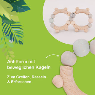 Holzspielzeug in Form einer Acht mit beweglichen Kugeln, geeignet zum Greifen, Rasseln und Erforschen. Im Hintergrund sind grüne Blätter sichtbar. Der Text auf dem Bild lautet: ‚Achtform mit beweglichen Kugeln. Zum Greifen, Rasseln & Erforschen.