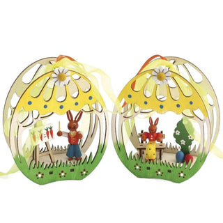 2 Fensterbilder aus Holz in 3D-Optik. Die Fensterbilder sind grün und gelb bemalt. In einem Bild befindet sich ein farbiger Osterhasenlehrer mit einer Schultafel und im zweiten Bild eine farbige Osterhäsin auf der Bank mit Ostereiern und einem Bäumchen