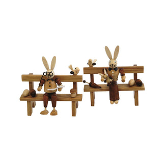 Naturfarbenes Osterhasenpaar. Der Osterhase und die Osterhäsin sitzen jeweils auf einer Bank. Der Hase liest gemütlich eine Zeitung und hält seine Brille in der Hand. Die Häsin hält ihr Hasenkind im Arm. Auf den Bänken befinden sich noch frühlingshafte Details wie Ostereier, Pilze und kleine Vögelchen