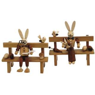 Naturfarbenes Osterhasenpaar. Der Osterhase und die Osterhäsin sitzen jeweils auf einer Bank. Der Hase liest gemütlich eine Zeitung und hält seine Brille in der Hand. Die Häsin hält ihr Hasenkind im Arm. Auf den Bänken befinden sich noch frühlingshafte Details wie Ostereier, Pilze und kleine Vögelchen