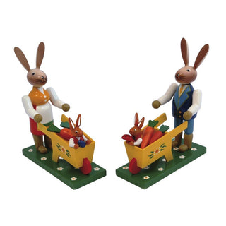 Farbiges Osterhasenpaar mit jeweils einer gelbfarbenen Schubkarre. In den Schubkarren sitzen die kleinen Hasenkinder mit Karotten und Ostereiern. Die Hasen befinden sich jeweils auf einem Sockel in der Optik einer Blumenwiese.
