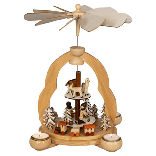 Ein Weihnachtspyramiden-Dekorationsstück aus Holz. Die Pyramide hat ein drehbares Flügelrad oben und zwei Ebenen mit kleinen Figuren. Auf der oberen Ebene befinden sich Rehe und ein Futterkrippe. Auf der unteren Ebene sind Tannenbäume und winterliche Figuren zu sehen. Vier Teelichte in runden Haltern sind um die Basis herum angeordnet und beleuchten die Szene sanft und sorgen für die nötige Thermik, um die Pyramide zum Drehen zu bewegen.