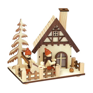 Das Bild zeigt ein Räucherhaus, das ein kleines Haus mit Schornstein, zwei Figuren mit roten Mützen und Schals, einen Baum und einen eingezäunten Hof darstellt. Eine Figur hält Skier, während die andere auf dem Schlitten sitzt. Das Haus weist detaillierte architektonische Elemente wie Fenster und Balken auf. Im Hof liegt auch ein kleiner Ball.