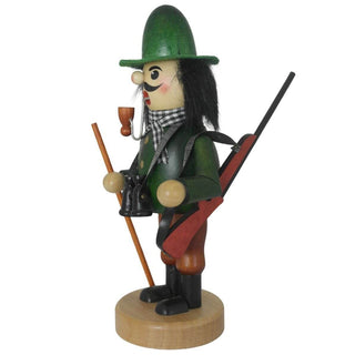 Ein traditioneller Räuchermann aus Holz steht auf einem runden Sockel. Er trägt grüne und braune Kleidung, einen grünen Hut, schwarze Stiefel und einen karierten Schal. Das Räuchermännchen hat ein Pfeifchen im Mund, hält einen Wanderstock und trägt ein Fernglas um den Hals. Über der Schulter trägt der Räuchermann "Jäger" ein Gewehr.