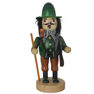 Ein traditioneller Räuchermann aus Holz steht auf einem runden Sockel. Er trägt grüne und braune Kleidung, einen grünen Hut, schwarze Stiefel und einen karierten Schal. Das Räuchermännchen hat ein Pfeifchen im Mund, hält einen Wanderstock und trägt ein Fernglas um den Hals. Über der Schulter trägt der Räuchermann "Jäger" ein Gewehr.