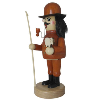Ein hölzerner Räuchermann in der Rolle eines Schäfers steht auf einem runden Sockel. Er trägt einen orangefarbenen Mantel mit zwei Knöpfen, schwarze Stiefel und einen braunen Hut. Der bärtige Mann hält ein kleines weißes Schaf in der linken Hand und einen langen Hirtenstab in der rechten. Seitenansicht