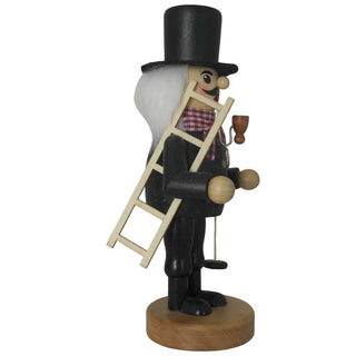 Ein Räuchermann "Schornsteinfeger" steht auf einem runden Sockel. Er trägt schwarze Kleidung mit goldenen Knöpfen, einen hohen schwarzen Hut, eine rot-weiß karierte Halsbinde und hat weißes Haar. In der Hand hält er einen Besen und ein Seil mit Gewicht, im Mund steckt eine Pfeife. Seitenansicht