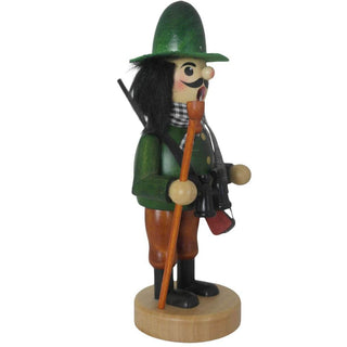 Ein traditioneller Räuchermann aus Holz steht auf einem runden Sockel. Er trägt grüne und braune Kleidung, einen grünen Hut, schwarze Stiefel und einen karierten Schal. Das Räuchermännchen hat ein Pfeifchen im Mund, hält einen Wanderstock und trägt ein Fernglas um den Hals. Über der Schulter trägt der Räuchermann "Jäger" ein Gewehr. Seitenansicht