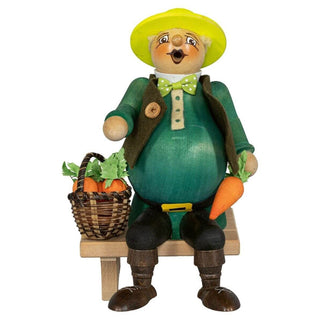 Das Bild zeigt ein sitzendes Räuchermännchen in Form eines dickbäuchigen Gärtners. Es trägt einen gelben Hut, eine grüne Jacke mit einem braunen Knopf und eine grüne Fliege mit weißen Punkten. In der rechten Hand hält es eine Karotte, während neben ihm ein Korb mit weiteren Karotten steht.