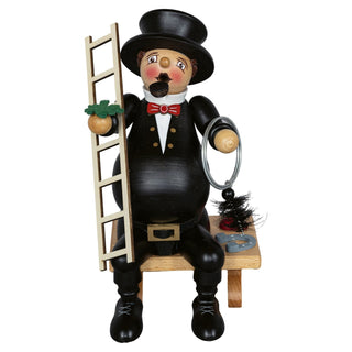 Das Bild zeigt ein Räuchermännchen in Form eines dickbauchigen Schornsteinfegers. Die Figur ist 23 cm groß und sitzt auf einer Bank. Der Schornsteinfeger trägt einen schwarzen Anzug mit goldenen Knöpfen, eine rote Fliege und einen schwarzen Zylinderhut. In der rechten Hand hält er eine Leiter, in der linken Hand ein Reinigungswerkzeug mit einem runden Bürstenkopf. Neben ihm auf der Bank liegen weitere Werkzeuge und ein Hufeisen.