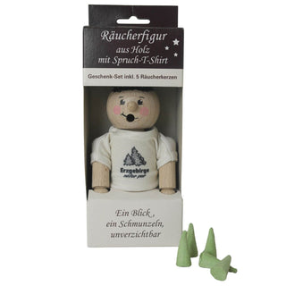 Eine hölzerne Räucherfigur mit einem weißen T-Shirt, auf dem "Erzgebirge natur pur" steht, im Geschenkkarton. Inklusive 5 Räucherkerzen. Auf der Verpackung steht: "Ein Blick, ein Schmunzeln, unverzichtbar". Fünf grüne Räucherkerzen sind vor dem Karton platziert.