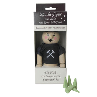 Ein hölzernes Räuchermännchen mit schwarzem T-Shirt, das gekreuzte Hämmer zeigt, in einer Box. Die Verpackung trägt die Aufschrift "Räucherfigur aus Holz mit Spruch-T-Shirt" und "Geschenk-Set inkl. 5 Räucherkerzen." Fünf grüne Räucherkerzen sind neben der Box platziert.