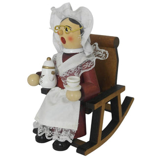 Ein hölzernes Räuchermännchen in Form einer älteren Frau sitzt auf einem Stuhl. Sie trägt traditionelle Kleidung mit weißer Haube, Brille und einer Schürze mit Spitzen. In der einen Hand hält sie eine Teekanne, in der anderen eine Teetasse. Ihr Gesicht zeigt rote Wangen. Seitenansicht