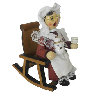 Ein hölzernes Räuchermännchen in Form einer älteren Frau sitzt auf einem Stuhl. Sie trägt traditionelle Kleidung mit weißer Haube, Brille und einer Schürze mit Spitzen. In der einen Hand hält sie eine Teekanne, in der anderen eine Teetasse. Ihr Gesicht zeigt rote Wangen. Seitenansicht