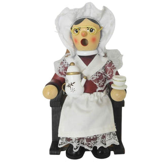 Ein hölzernes Räuchermännchen in Form einer älteren Frau sitzt auf einem Stuhl. Sie trägt traditionelle Kleidung mit weißer Haube, Brille und einer Schürze mit Spitzen. In der einen Hand hält sie eine Teekanne, in der anderen eine Teetasse. Ihr Gesicht zeigt rote Wangen. 