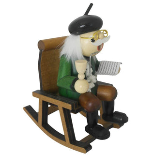 Ein hölzernes Räuchermännchen in Form eines älteren Mannes sitzt auf einem Holzstuhl. Er trägt grüne Kleidung, braune Hose, schwarze Schuhe, eine Brille und einen schwarzen Hut. In der einen Hand hält er eine Pfeife, in der anderen eine Zeitung. Seitenansicht