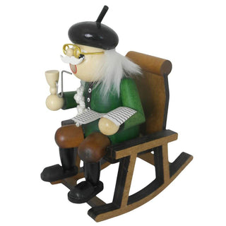 Ein hölzernes Räuchermännchen in Form eines älteren Mannes sitzt auf einem Holzstuhl. Er trägt grüne Kleidung, braune Hose, schwarze Schuhe, eine Brille und einen schwarzen Hut. In der einen Hand hält er eine Pfeife, in der anderen eine Zeitung. Seitenansicht