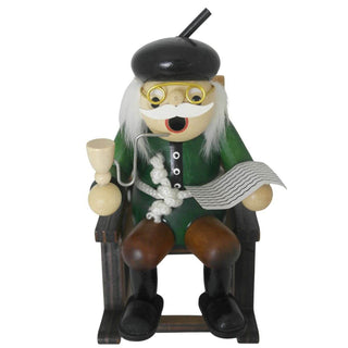 Ein hölzernes Räuchermännchen in Form eines älteren Mannes sitzt auf einem Holzstuhl. Er trägt grüne Kleidung, braune Hose, schwarze Schuhe, eine Brille und einen schwarzen Hut. In der einen Hand hält er eine Pfeife, in der anderen eine Zeitung.