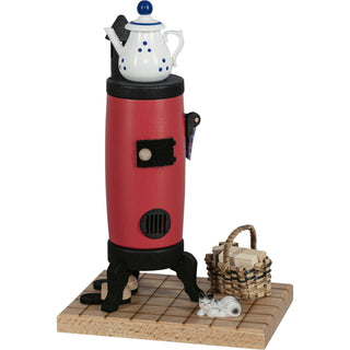 Räucherofen in Form eines roten Küchenherds mit schwarzem Sockel und Füßen, eine kleine weiße Teekanne mit blauen Punkten steht oben auf dem Ofen, mit einem kleinen Korb mit Holzscheiten und einer grauen Katze davor liegend, auf einem Holzboden. Pantoffeln stehen am Ofen.