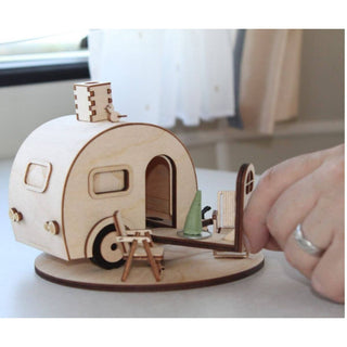 Das Bild zeigt ein kleines, detailliertes Holzmodell eines Wohnwagens. Das Modell hat Fenster, eine Tür und einen Schornstein mit einem Vogel darauf. Es gibt auch eine Außenszene mit Miniaturmöbeln, darunter Stühle und ein Tisch mit einem Gegenstand darauf. Eine Hand ist im Bild zu sehen, die mit dem Modell interagiert.