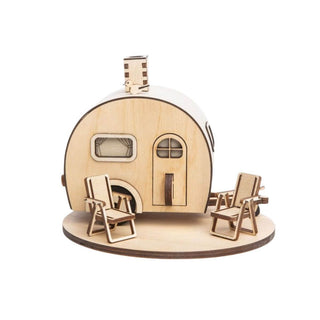 Das Bild zeigt ein kleines, hölzernes Modell eines Wohnwagens. Das Modell steht auf einer runden Holzplatte und hat zwei Holzstühle vor dem Wohnwagen. Der Wohnwagen hat ein kleines rechteckiges Fenster mit Vorhängen und eine gewölbte Tür mit Griff. Auf dem Dach des Wohnwagens befindet sich ein Schornstein.