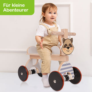 Ein kleines Kind sitzt auf einem hölzernen Laufrad mit vier Rädern, einem Lenker und einer Frontplatte mit einem Waschbärgesicht. Das Kind trägt einen weißen Kurzarm-Body und eine beige Latzhose. Oben links im Bild steht in einem grünen Feld der Text: „Für kleine Abenteurer“.