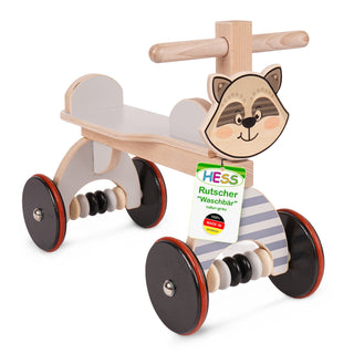 Ein hölzernes Rutschfahrzeug für Kinder mit vier schwarzen Rädern und roten Felgen. Es hat einen Sitz und Lenker zum Festhalten. Auf der Vorderseite ist ein Waschbärgesicht abgebildet. Ein Etikett an der Vorderseite trägt die Aufschrift: „HESS Rutscher 'Waschbär' natur grau 100% MADE IN GERMANY“.