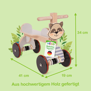 Ein hölzernes Kinder-Rutschfahrzeug in Form eines Waschbären mit vier schwarzen Rädern und roten Akzenten. Es verfügt über eine Sitzfläche und einen Griff zum Festhalten. Auf der Vorderseite ist ein Waschbärgesicht abgebildet. Ein Schild am Fahrzeug zeigt die Aufschrift: „HESS Rutscher 'Waschbär' Art.Nr.: 31179“. Unten im Bild steht: „Aus hochwertigem Holz gefertigt“. Die Maße des Fahrzeugs sind 41 cm lang, 19 cm breit und 34 cm hoch.