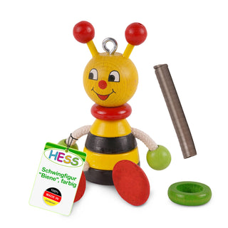 Ein buntes Holzspielzeug in Form einer Biene mit gelbem Kopf, roten Fühlern, schwarzen und weißen Augen, roter Nase und Mund. Der Körper ist gelb-schwarz gestreift, mit roten Füßen und grünen Händen, die an weißen Seilarmen befestigt sind. Neben der Figur liegen eine Metallfeder, ein grüner Ring und ein Schild mit der Aufschrift: „HESS Schwingfigur 'Biene', farbig 100% MADE IN GERMANY“.