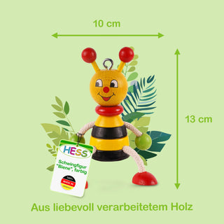  Eine bunte Schwingfigur in Form einer Biene aus Holz, etwa 13 cm hoch und 10 cm breit. Die Biene hat einen gelben Kopf mit roten Fühlern und rotem Mund sowie einen schwarz-gelb gestreiften Körper. Ihre Beine bestehen aus weißen Seilen und enden in roten und grünen Kugeln. An der linken Hand hängt ein Schild mit der Aufschrift: „HESS Schwingfigur 'Biene', farbig“ und „100% Made in Germany“. Im Hintergrund sind stilisierte grüne Blätter zu sehen. Am unteren Bildrand steht: „Aus liebevoll verarbeitetem Holz“.