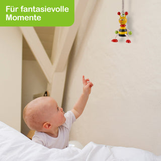 Ein Baby liegt in einem Bett und zeigt mit dem Finger auf ein buntes Spielzeug in Form einer Biene, das an der Decke hängt. Die Biene ist rot, grün und gelb gefärbt. In der oberen linken Ecke befindet sich ein grünes Textfeld mit der Aufschrift: „Für fantasievolle Momente“.