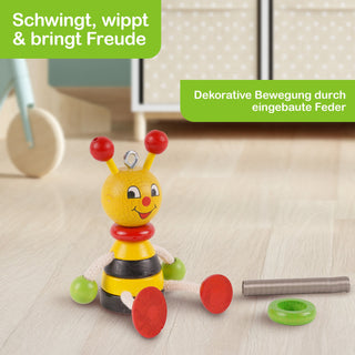 Bunte Holz-Biene mit freundlichem Gesicht sitzt auf hellem Boden. Arme und Beine aus Seil mit roten und grünen Kugeln. Im Hintergrund Möbel, daneben Metallstab und grüner Ring. Text im Bild: „Schwingt, wippt & bringt Freude“ und „Dekorative Bewegung durch eingebaute Feder“.grüner Ring. Im Hintergrund sind unscharfe Möbel zu sehen. Oben links steht in einem grünen Kasten: „Schwingt, wippt & bringt Freude“, oben rechts: „Dekorative B