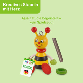 Eine bunte Holzfigur in Form einer Biene mit gelbem Kopf, roten Fühlern, freundlichem Gesicht und gelb-schwarz gestreiftem Körper. Daneben liegen rote und grüne Ringe sowie ein Etikett mit der Aufschrift: „Hess Schwingfigur 'Biene', farbig“. Der Hintergrund ist hellgrün. Oben links steht in einem grünen Feld: „Kreatives Stapeln mit Herz“, darunter der Text: „Qualität, die begeistert – kein Spielzeug!“.