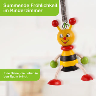 Ein fröhlich lächelndes Holzspielzeug in Form einer Biene hängt an einer Metallfeder. Die Biene hat einen gelben Kopf mit roten Fühlern, einen rot-gelb-schwarz gestreiften Körper, rote Füße sowie weiße Seilarme und -beine mit grünen Händen. Der Hintergrund zeigt einen unscharfen Raum. Oben links steht in weißer Schrift auf grünem Hintergrund: „Summende Fröhlichkeit im Kinderzimmer“, unten links: „Eine Biene, die Leben in den Raum bringt“.