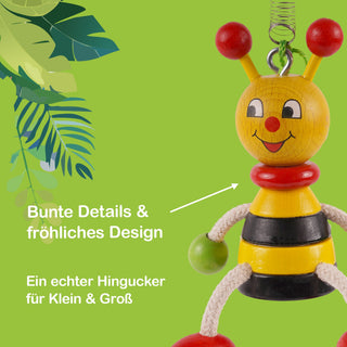 Ein fröhlich gestaltetes Holzspielzeug in Form einer Biene mit gelbem Kopf, roten Fühlern und einem freundlichen Gesicht. Der Körper ist schwarz-gelb gestreift, die Arme bestehen aus weißen Seilen mit grünen Kugeln an den Enden. Im Hintergrund sind grüne Blätter zu sehen. Im Bild steht der Text: „Bunte Details & fröhliches Design“ und „Ein echter Hingucker für Klein & Groß“.