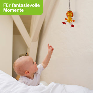 Ein Baby liegt in einem Bett und zeigt mit dem Finger auf ein hängendes Spielzeug in Löwenform. Oben links im Bild steht in einem grünen Feld: „Für fantasievolle Momente“.