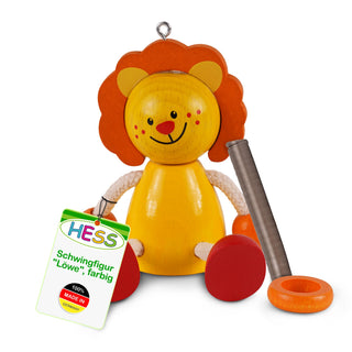 Eine bunte Holzfigur in Form eines Löwen mit gelbem Körper, orangefarbenem Kopf und Mähne sowie roten Füßen. Der Löwe hat ein freundliches Gesicht mit schwarzen Augen, roter Nase und lächelndem Mund. An der linken Seite hängt ein Etikett mit der Aufschrift: „HESS Schwingfigur 'Löwe', farbig – 100% MADE IN GERMANY“. Neben der Figur liegen eine Metallfeder und ein orangefarbener Ring.
