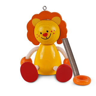 Ein buntes Holzspielzeug in Form eines sitzenden Löwen mit gelbem Körper, orangefarbenem Gesicht und Mähne sowie roten Füßen. Der Löwe hat bewegliche Arme aus weißem Seil und hält einen metallischen Zylinder in der rechten Hand. Vor ihm liegt ein orangefarbener Ring.