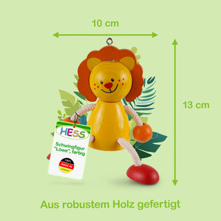 Ein farbiges Holzspielzeug in Form eines Löwen, etwa 13 cm hoch und 10 cm breit. Der Löwe hat eine orangefarbene Mähne, gelbes Gesicht mit roten Wangen und rotem Mund. Arme und Beine bestehen aus weißen Seilen mit orangefarbenen Kugeln und roten Füßen. Ein Etikett trägt die Aufschrift: „HESS Schwingfigur 'Löwe', farbig – 100% MADE IN GERMANY“. Im Hintergrund sind grüne Blätter zu sehen. Unten im Bild steht: „Aus robustem Holz gefertigt“.