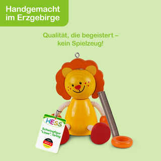 Eine handgefertigte Holzfigur in Form eines Löwen mit gelbem Körper, orangefarbener Mähne und roten Füßen. Die Figur hält ein Schild mit der Aufschrift: „Hess Schwingfigur 'Löwe', farbig“. Oben links im Bild steht: „Handgemacht im Erzgebirge“, in der Mitte: „Qualität, die begeistert – kein Spielzeug!“.