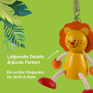  Ein bunter Holzlöwe mit lächelndem Gesicht hängt an einer Metallfeder. Der Hintergrund ist grün mit illustrierten Blättern auf der linken Seite. Im Bild steht der Text: „Liebevolle Details & bunte Farben“ und „Ein echter Hingucker für Groß & Klein“.