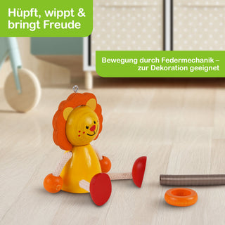 Ein buntes Holzspielzeug in Form eines sitzenden Löwen mit orangefarbener Mähne und roten Füßen steht auf einem hellen Holzboden. Der Löwe hat ein fröhliches Gesicht. Daneben liegen eine Federmechanik und ein orangefarbener Ring. Im Hintergrund sind unscharfe Möbel zu sehen. Im Bild stehen die Texte: „Hüpft, wippt & bringt Freude“ und „Bewegung durch Federmechanik – zur Dekoration geeignet“.