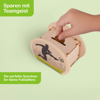 Rechteckige Spardose aus Holz mit abgerundeten Ecken. Auf der Vorderseite ist ein schwarzer Fußballspieler zu sehen, der einen Ball tritt, vor einem Hintergrund mit jubelnden Fans und grünem Rasen. Oben befindet sich ein Münzschlitz, über dem eine Hand eine Münze hält. Der Hintergrund ist hellgrün. Auf der Spardose steht: „Sparen mit Teamgeist“ und „Die perfekte Spardose für kleine Fußballfans“.