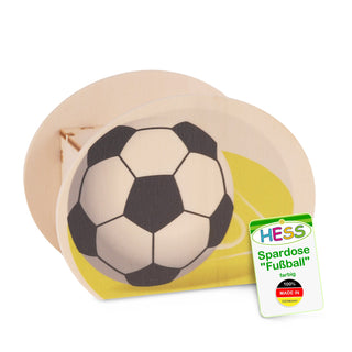 Ovale Spardose aus Holz mit einem schwarz-weißen Fußballmotiv auf der Vorderseite. Die Spardose steht auf einer gelben Fläche. Unten rechts befindet sich ein Etikett mit der Aufschrift: „HESS Spardose 'Fußball' farbig 100% MADE IN GERMANY“.