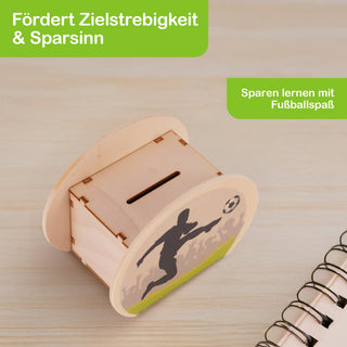 Rechteckige Spardose aus hellem Holz mit abgerundeten Seiten. Auf einer Seite ist ein Fußballspieler abgebildet, der einen Ball kickt. Im Hintergrund sind stilisierte Zuschauer in Grautönen und ein grüner Streifen am unteren Rand zu sehen. Oben befindet sich ein Schlitz zum Einwerfen von Münzen. Auf der Spardose steht: „Fördert Zielstrebigkeit & Sparsinn“ und „Sparen lernen mit Fußballspaß“.