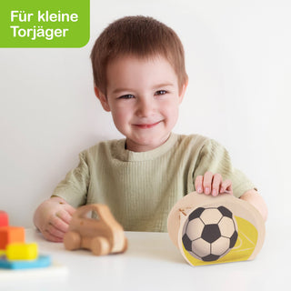 Runde Spardose in Form eines klassischen schwarz-weißen Fußballs. Sie wird von einem Kind gehalten, das an einem Tisch sitzt. Im Hintergrund sind ein gelber Untergrund, bunte Bauklötze und ein kleines Holzauto zu sehen. Oben links steht in weißer Schrift auf grünem Hintergrund: „Für kleine Torjäger“.