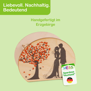 Ovale Spardose aus hellem Holz mit einem Motiv auf der Vorderseite: Eine schwarze Silhouette eines Brautpaares steht unter einem Baum mit roten Blättern. Oben links im Bild steht in weißer Schrift auf grünem Hintergrund: „Liebevoll. Nachhaltig. Bedeutend“. Darunter in grüner Schrift: „Handgefertigt im Erzgebirge“. Unten rechts befindet sich ein Etikett mit der Aufschrift: „HESS Spardose 'Hochzeit' 100% MADE IN GERMANY“.